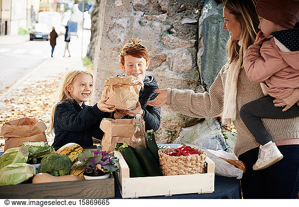 Lächelnde männliche und weibliche Geschwister kaufen Gemüse von weiblichen Marktverkäufern