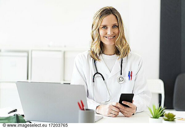 Lächelnde medizinische Mitarbeiterin mit Smartphone und Laptop am Schreibtisch in einer medizinischen Klinik