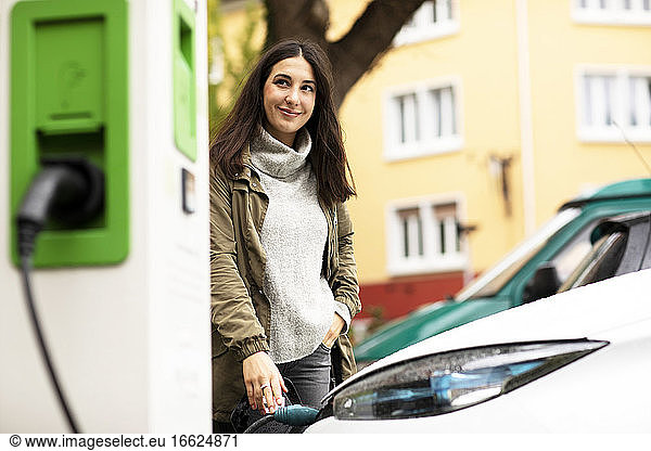 Lächelnde schöne Frau  die wegschaut  während sie mit der Hand in der Tasche steht und ein Elektrofahrzeug an einer Station auflädt