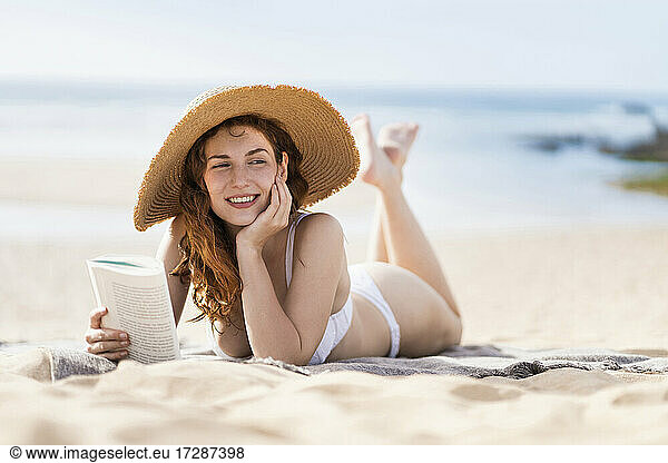 Lächelnde schöne Frau mit Buch Tag träumt  während auf Sand während sonnigen Tag liegen