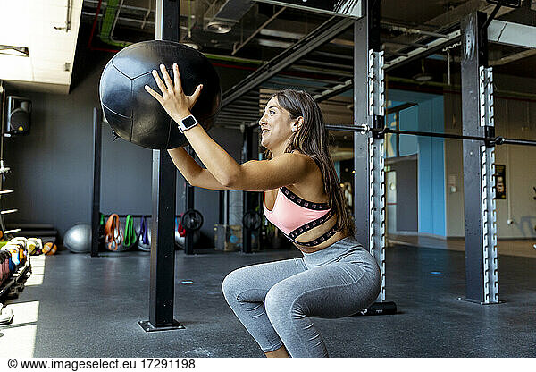 Lächelnde Sportlerin hockt beim Training mit einem Fitnessball im Fitnessstudio