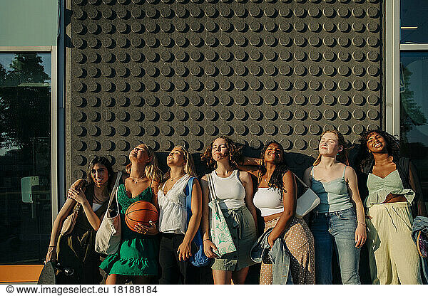 Lächelnde Teenager-Mädchen mit geschlossenen Augen genießen den sonnigen Tag  während sie an der Wand stehen