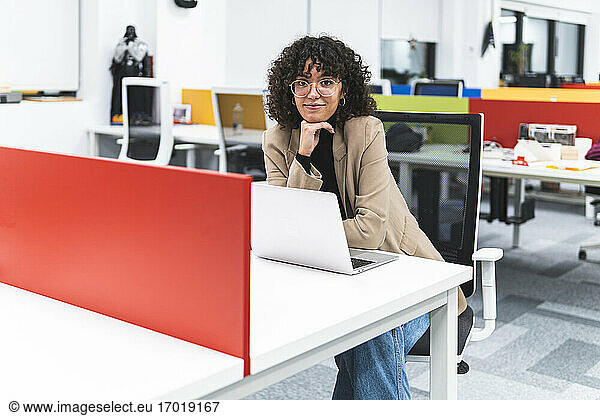 Lächelnde weibliche Fachkraft mit Hand am Kinn am Schreibtisch am Arbeitsplatz