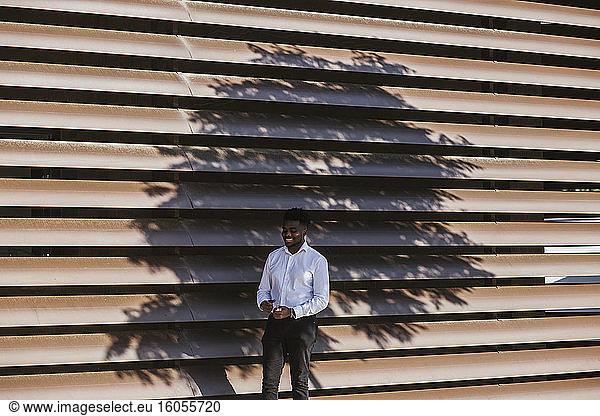 Lächelnder Geschäftsmann  der vor einem modernen Bauwerk mit Baumschatten steht