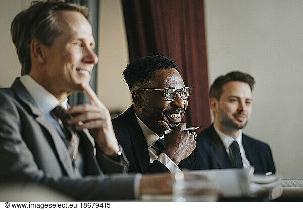 Lächelnder Geschäftsmann mit Hand am Kinn  der mit männlichen Kollegen während einer Besprechung im Büro sitzt