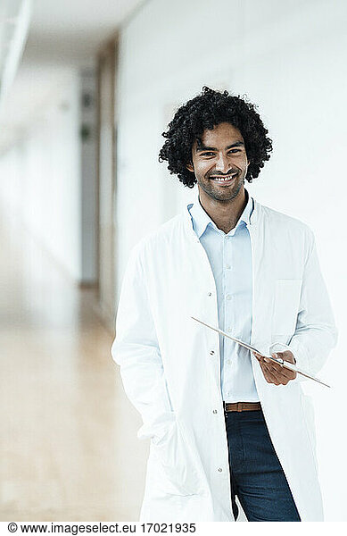 Lächelnder junger Mann im Gesundheitswesen  der ein Klemmbrett hält  während er im Krankenhausflur steht