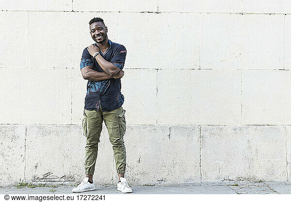 Lächelnder junger Mann in Freizeitkleidung an einer weißen Wand stehend