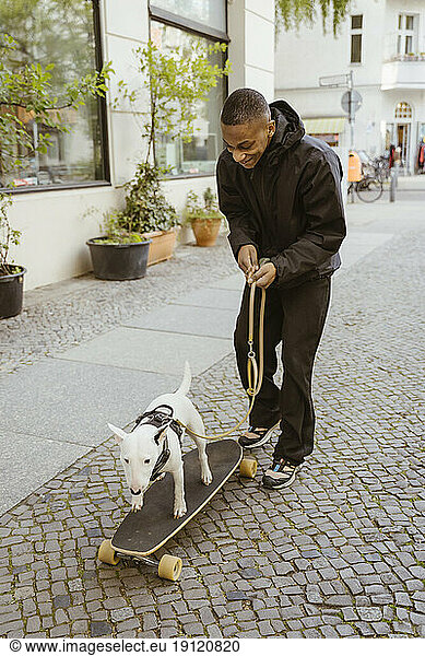 Lächelnder junger Mann in voller Länge mit Bullterrier auf Skateboard auf dem Bürgersteig