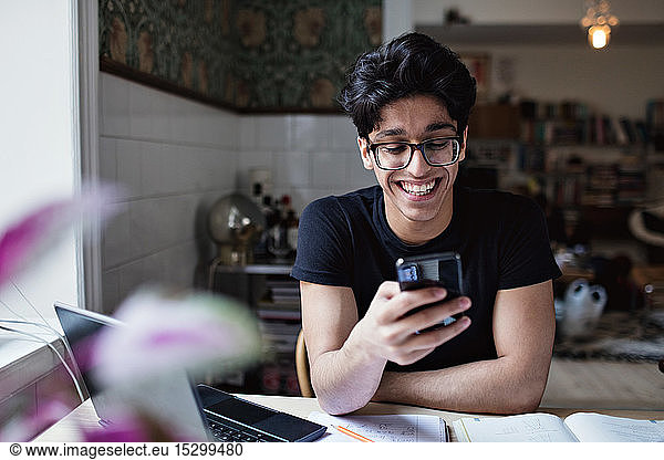 Lächelnder junger Mann nutzt soziale Medien  während er zu Hause Hausaufgaben macht