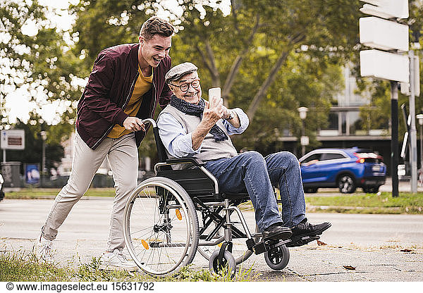 Lächelnder junger Mann schiebt glücklichen älteren Mann mit Smartphone im Rollstuhl
