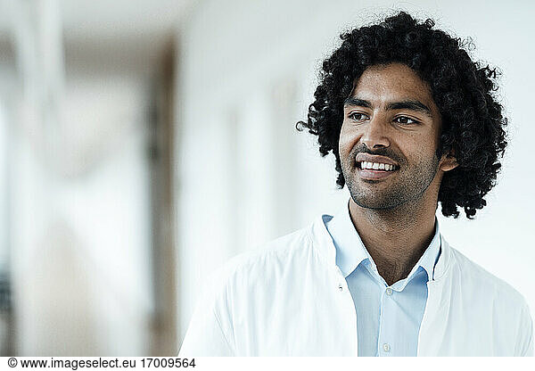 Lächelnder männlicher Angestellter im Gesundheitswesen mit schwarzen lockigen Haaren  der ins Krankenhaus blickt