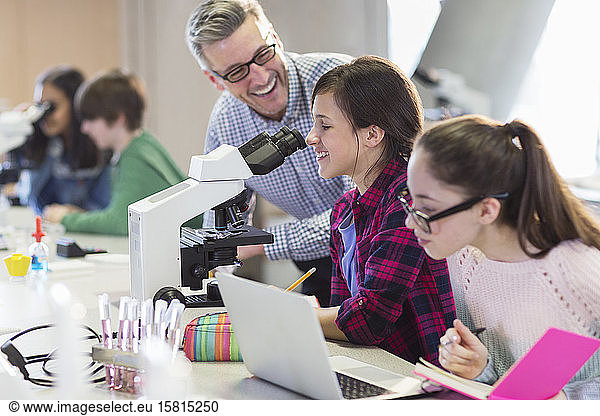 Lächelnder männlicher Lehrer der Naturwissenschaften hilft Schülerinnen bei der Durchführung eines wissenschaftlichen Experiments am Mikroskop im Labor
