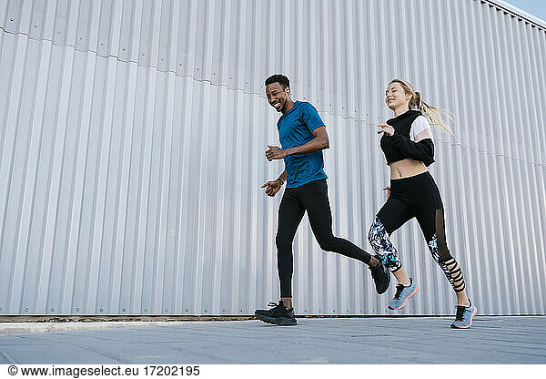 Lächelnder männlicher und weiblicher Athlet  der während des Sporttrainings auf dem Gehweg an einer Mauer läuft