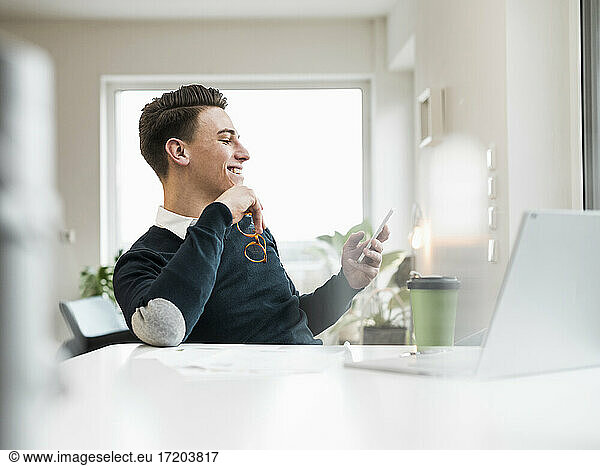 Lächelnder männlicher Unternehmer  der im Büro zu Hause sitzt und über sein Smartphone im Internet surft