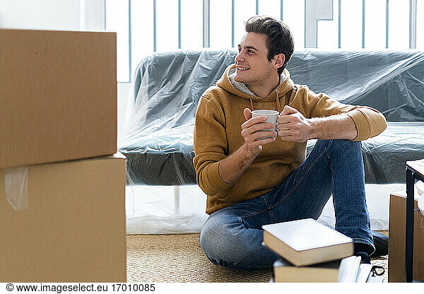 Lächelnder Mann mit Kaffeetasse  der beim Auspacken in der neuen Wohnung wegschaut