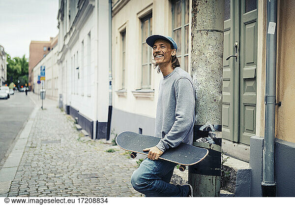 Lächelnder Mann mit Skateboard lehnt an Mast in der Stadt