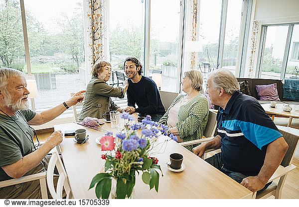 Lächelnder Mann sitzt inmitten älterer Menschen  die am Esstisch gegen das Fenster im Pflegeheim sitzen