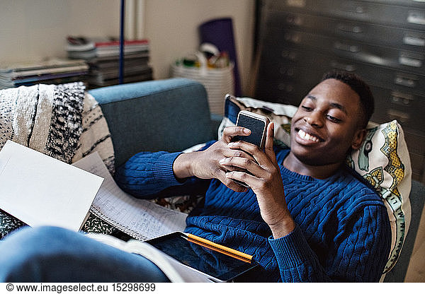 Lächelnder Teenager nutzt soziale Medien  während er bei den Hausaufgaben im Wohnzimmer auf dem Sofa liegt
