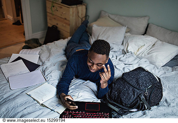 Lächelnder Teenager nutzt soziale Medien  während er zu Hause im Bett seine Hausaufgaben macht