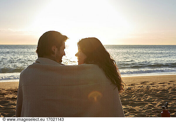 Lächelndes  in eine Decke gehülltes Paar sieht sich am Strand sitzend an