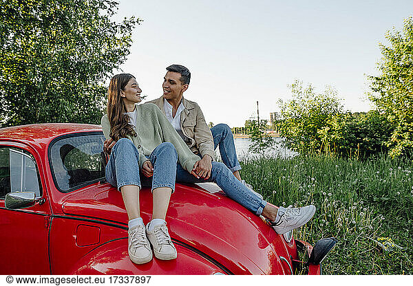 Lächelndes junges Paar  das sich auf der Motorhaube eines roten Autos sitzend ansieht