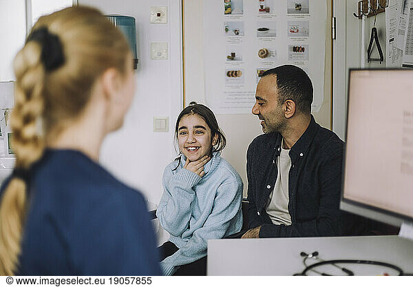 Lächelndes Mädchen erklärt dem Arzt  während es mit seinem Vater bei einem Besuch in der Klinik sitzt