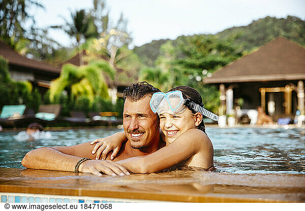 Lächelndes Mädchen mit Arm um Vater im Schwimmbad am Urlaubsort