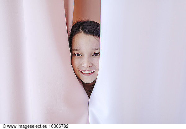 Lächelndes Mädchen versteckt sich hinter einem Vorhang zu Hause