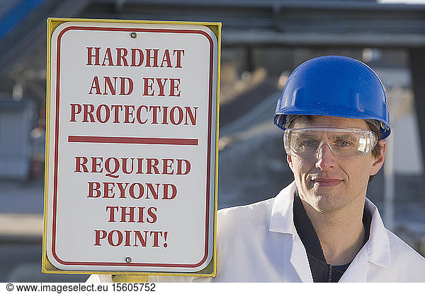 Lab worker beside a warning sign saying 'Hardhat and Eye Protection Required Beyond This Point' at an asphalt plant
