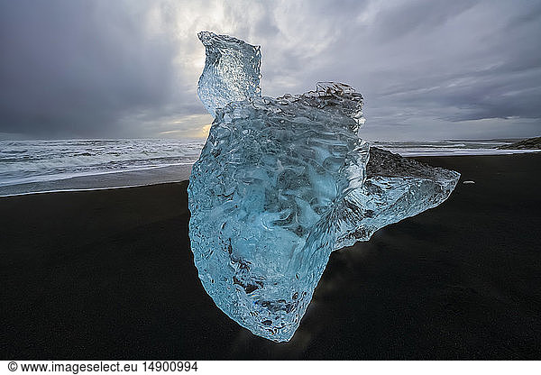 Large chunk of ice sitting on the shore of Iceland with dramatic skies in behind it; Iceland
