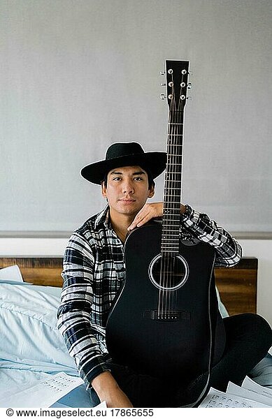 Latin man sitting on bed holding a guitar