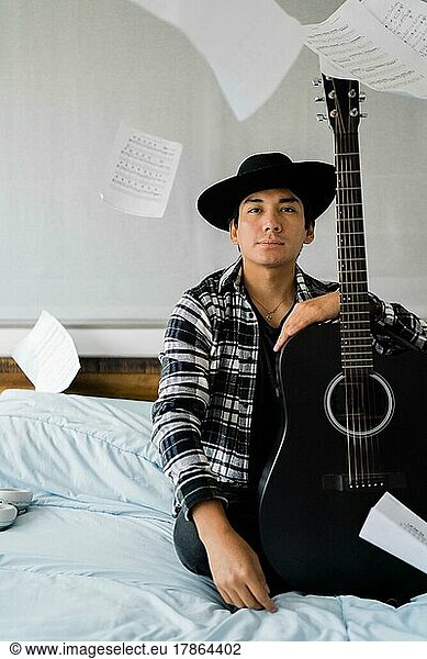 Latin man sitting on bed with guitar and note sheets floating around
