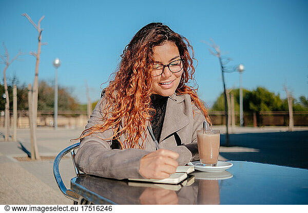 latin woman drinks coffee and writes notes  on a metal table outdoors