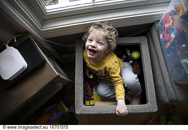 Laughing Boy Sits in Toy Box Next to Storage Bins Looks Up at Camera