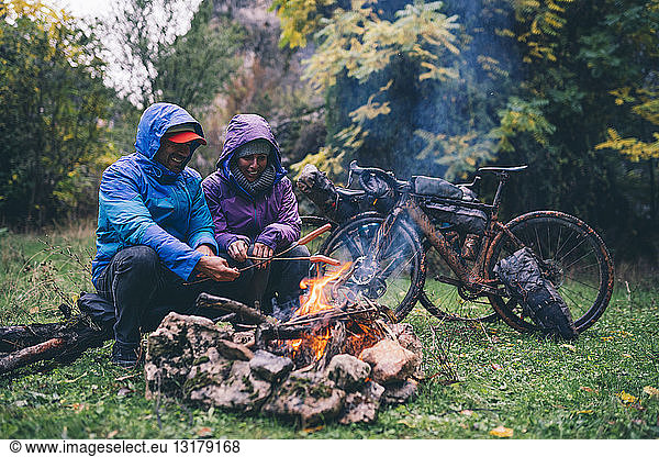 Laughing couple with with bmx bikes sitting at camp fire grilling sausages