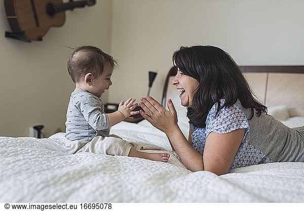 Laughing mid-30's mom clapping hands on bed with 1 yr old baby
