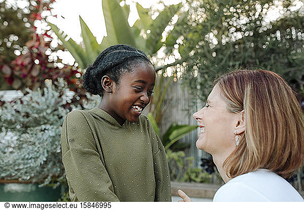 Laughing white mom and black daughter in front of lush greenery