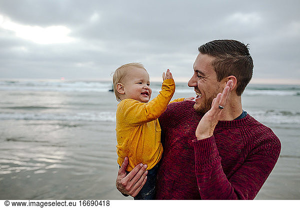 Laughing 30 yr old father holding baby at the ocean giving high-fives