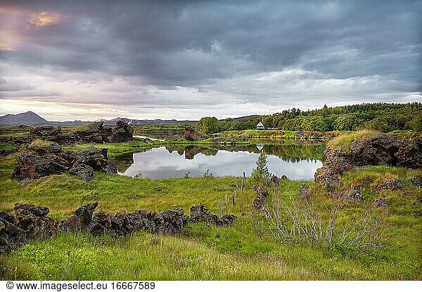 Lavaformationen aus vulkanischem Gestein ragen aus grüner Wiese mit dramatischem Himmel am Kalfaströnd  Haus spiegelt sich im See Mývatn  Skútustaðir  Norðurland eystra  Island  Europa