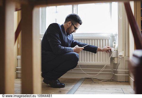 Lawyer crouching while charging phone by radiator with furniture in foreground