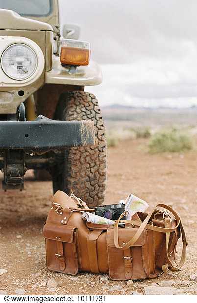 Leather overnight bag with a camera and map standing next to the wheel of a 4x4 parked in the desert.