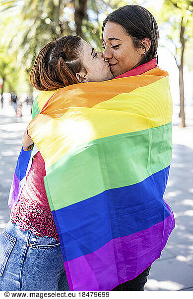 Lesbian couple covered in multi colored flag kissing on mouth at footpath