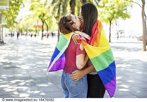 Lesbian couple covered in rainbow colored flag hugging each other on footpath