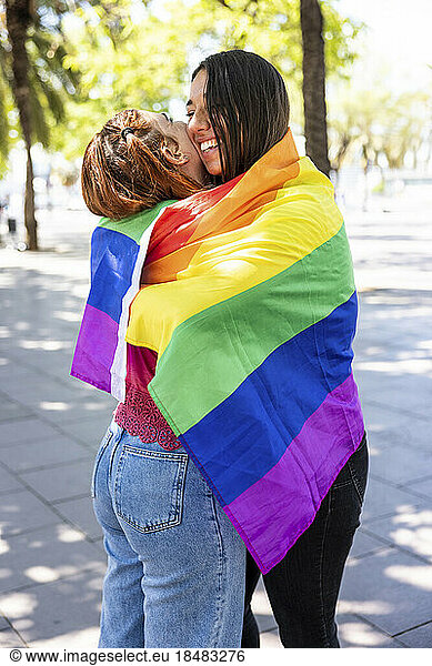 Lesbian couple hugging inside rainbow colored flag on footpath
