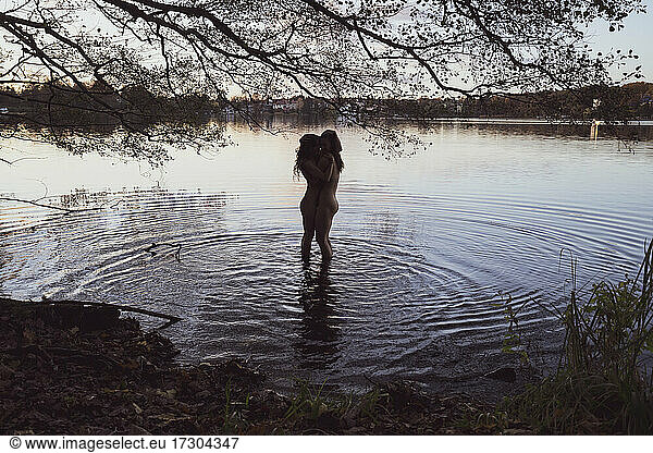 lesbian queer couple hug nude in rippling lake water under tree