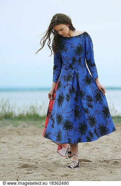Lifestyle portrait of a young woman n blue dress with prints walking on the beach