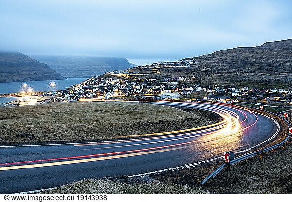 LIght trails on road with Eidi town in background at dusk  Faroe Islands