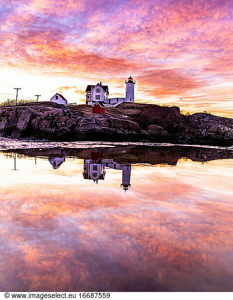 Lighthouse and colorful pink clouds reflecting in puddle at sunrise.