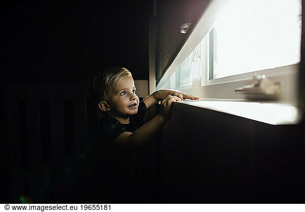 Little blonde boy looks outside of dark room through bright wind
