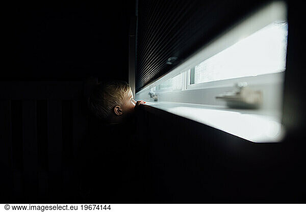 Little blonde boy peaks out of dark room through bright window